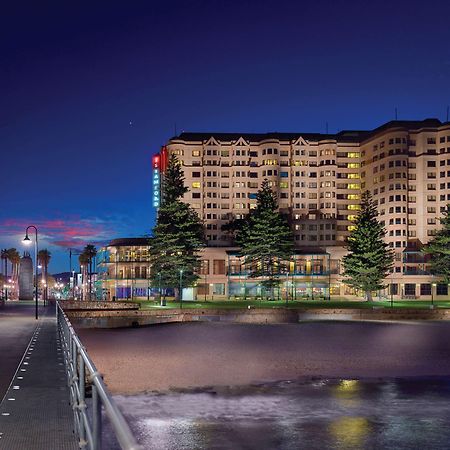 Hotel Stamford Grand Adelaide Glenelg Zewnętrze zdjęcie The photo shows a waterfront scene at dusk, featuring a large building lit up against a darkening sky. The architecture appears modern and prominent, with several windows illuminated. In the foreground, there is a walkway or pier with fencing along t