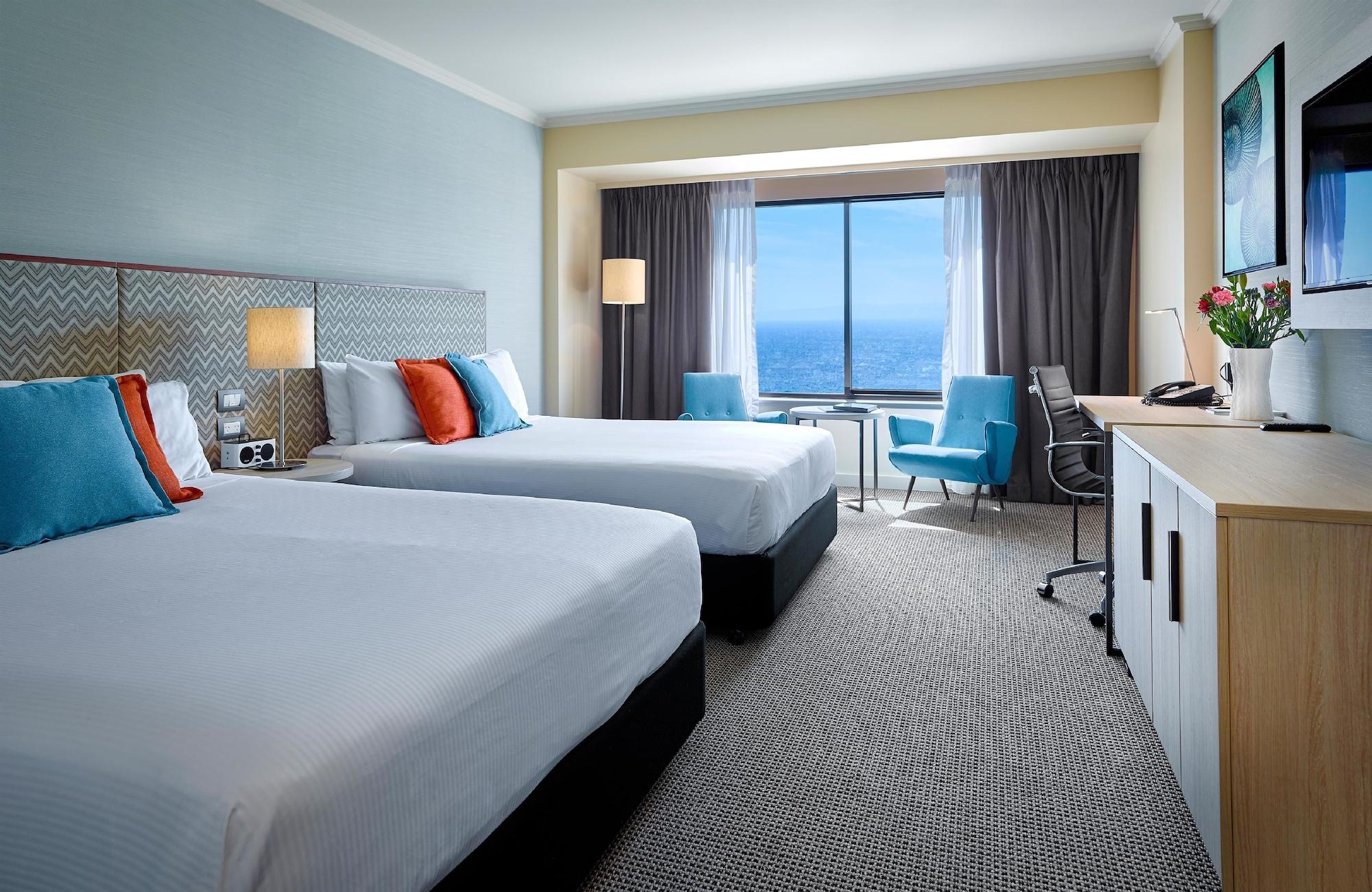 Hotel Stamford Grand Adelaide Glenelg Zewnętrze zdjęcie The photo shows a well-decorated hotel room featuring two queen-sized beds with white linens and colorful accent pillows. There is a large window with curtains that offers a view of the ocean. A small seating area with a blue chair and a matching des