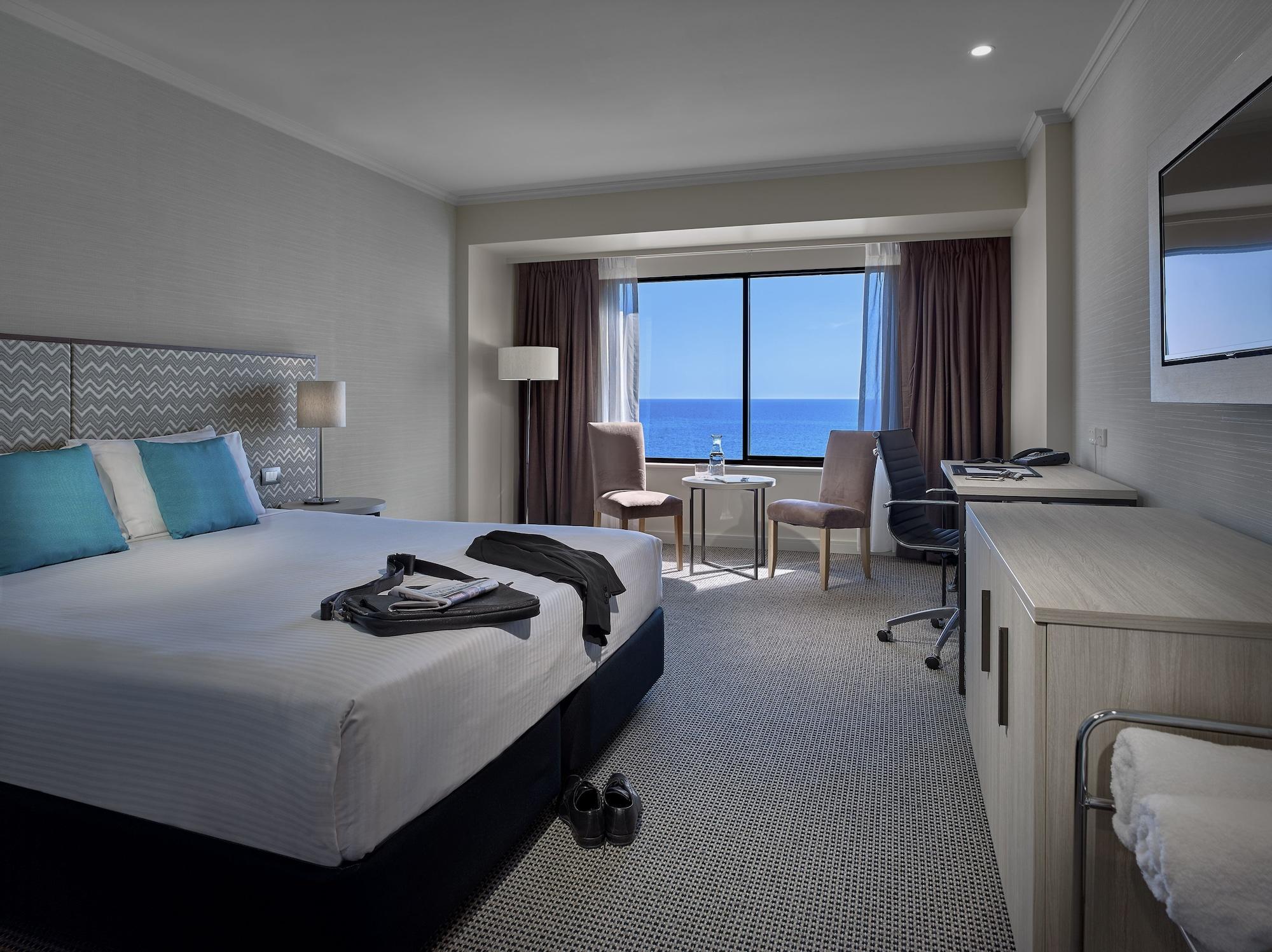Hotel Stamford Grand Adelaide Glenelg Zewnętrze zdjęcie The photo shows a modern hotel room featuring a queen-sized bed with white linens and two aqua blue decorative pillows. There are bedside lamps and a large window with a view of the ocean. The room also has a small seating area with two chairs and a 