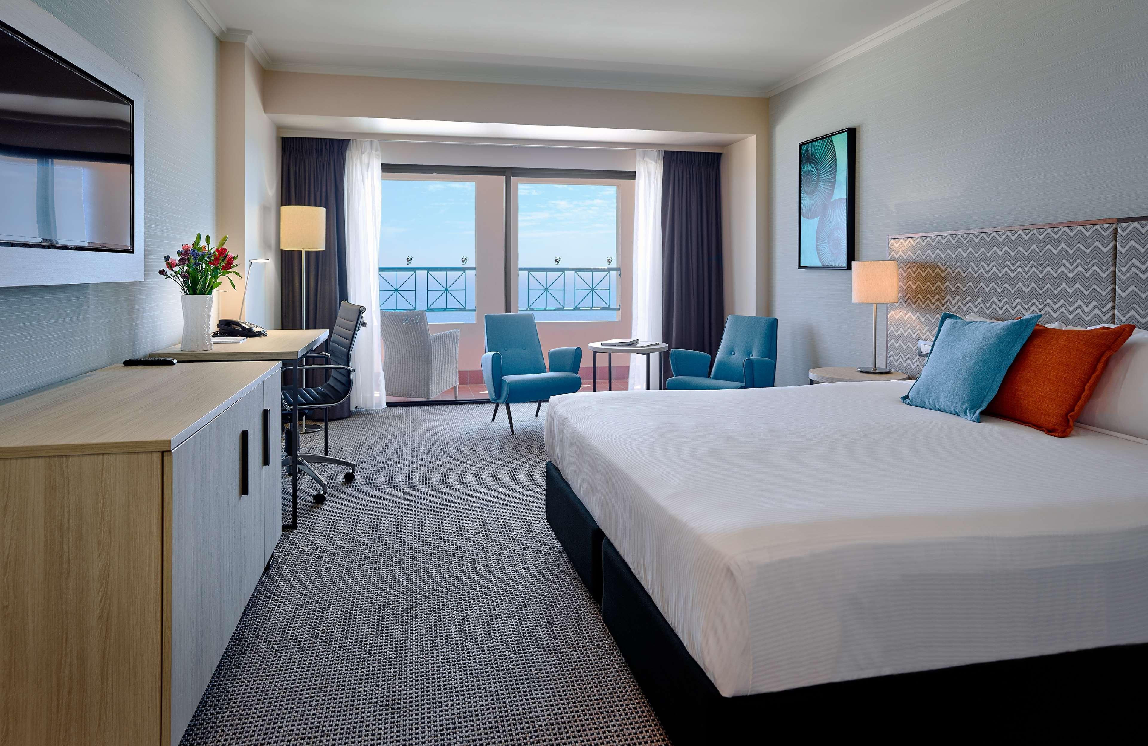 Hotel Stamford Grand Adelaide Glenelg Zewnętrze zdjęcie The photo depicts a modern hotel room. The room features a large bed with a white comforter, and there are decorative pillows in orange and blue. There is a flat-screen TV mounted on the wall, and a desk with an office chair is positioned near the wi