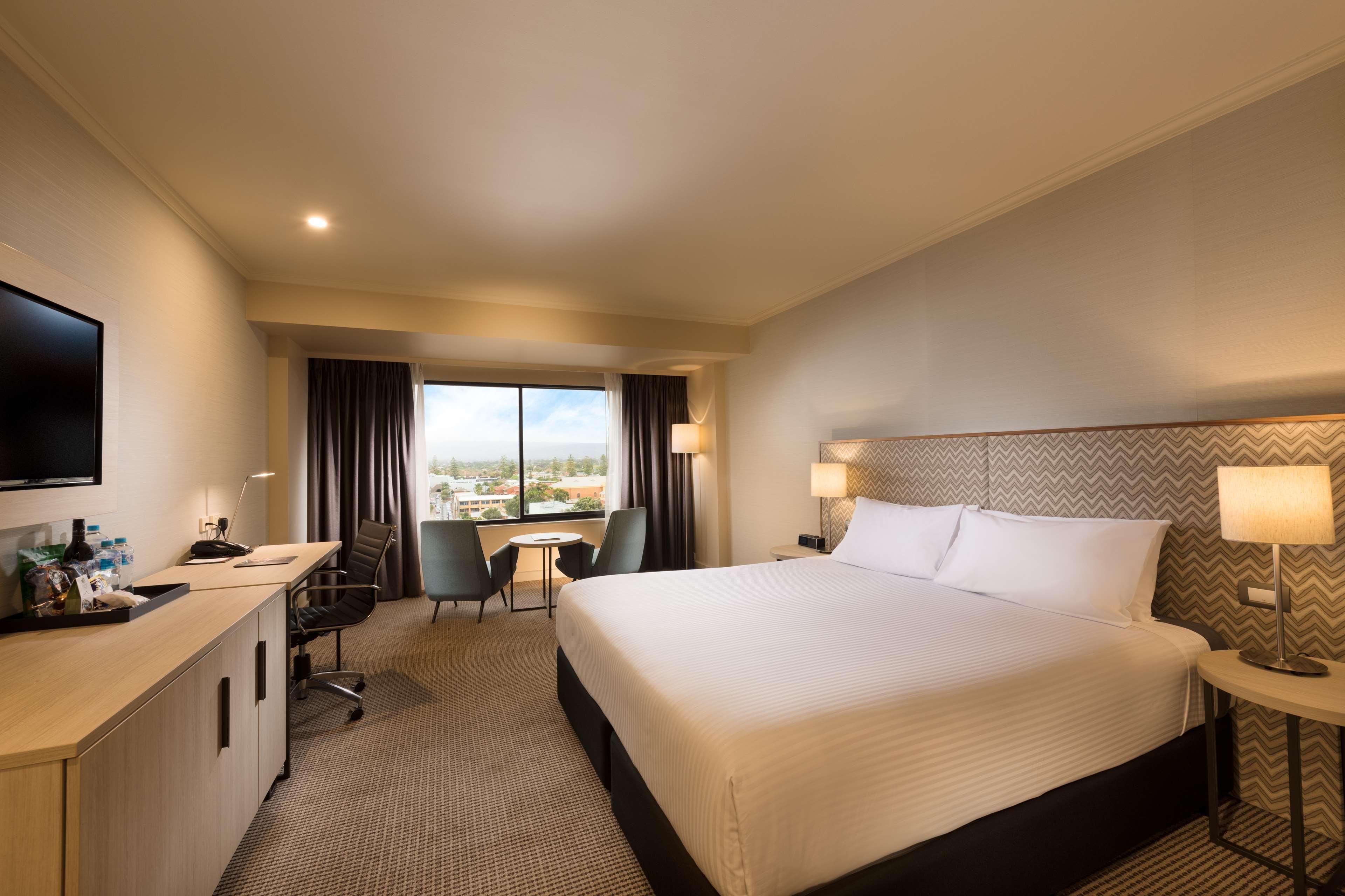 Hotel Stamford Grand Adelaide Glenelg Zewnętrze zdjęcie The photo shows a modern hotel room. It features a large bed with white linens, a desk with a chair, and a flat-screen TV mounted on the wall. There are two armchairs and a small table near a large window that lets in natural light, providing a view 