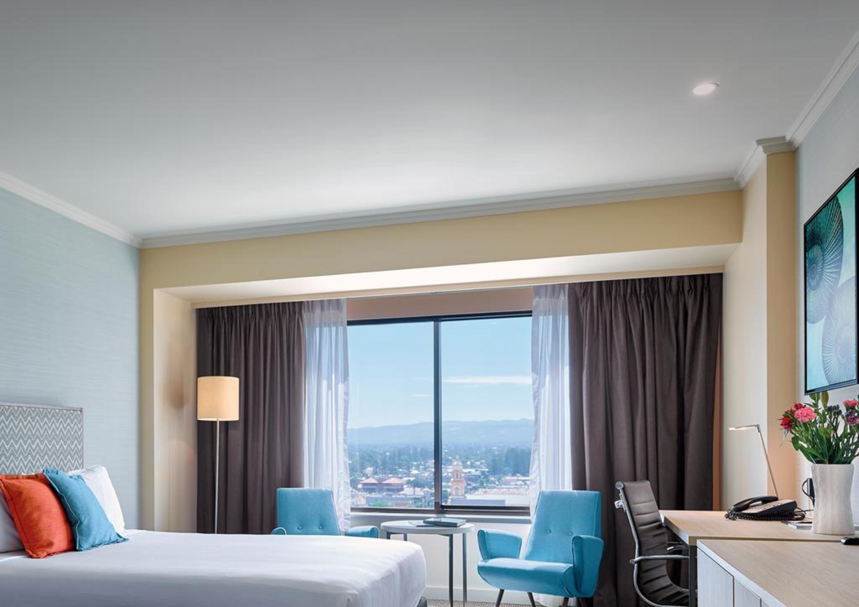 Hotel Stamford Grand Adelaide Glenelg Zewnętrze zdjęcie The photo shows a modern hotel room featuring a queen-sized bed with white linens and colorful decorative pillows. There are two blue chairs beside a small table positioned near a large window that provides a view of the outside, with sheer and dark 
