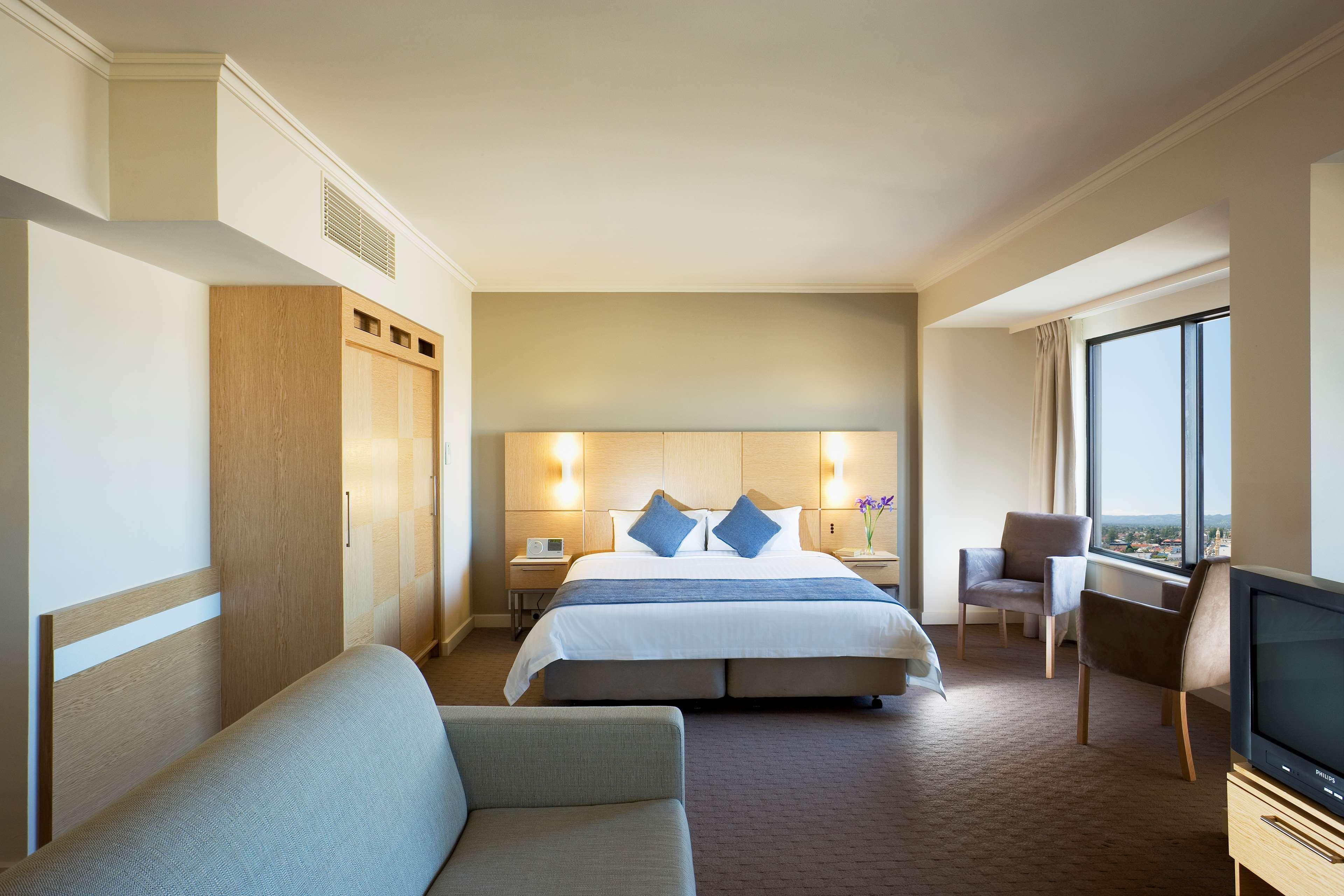 Hotel Stamford Grand Adelaide Glenelg Pokój zdjęcie The photo shows a modern hotel room featuring a large bed with two blue decorative pillows. There are bedside lamps on both sides of the headboard. To the left, there's a wooden wardrobe and a small table with a vase and a few amenities. On the right