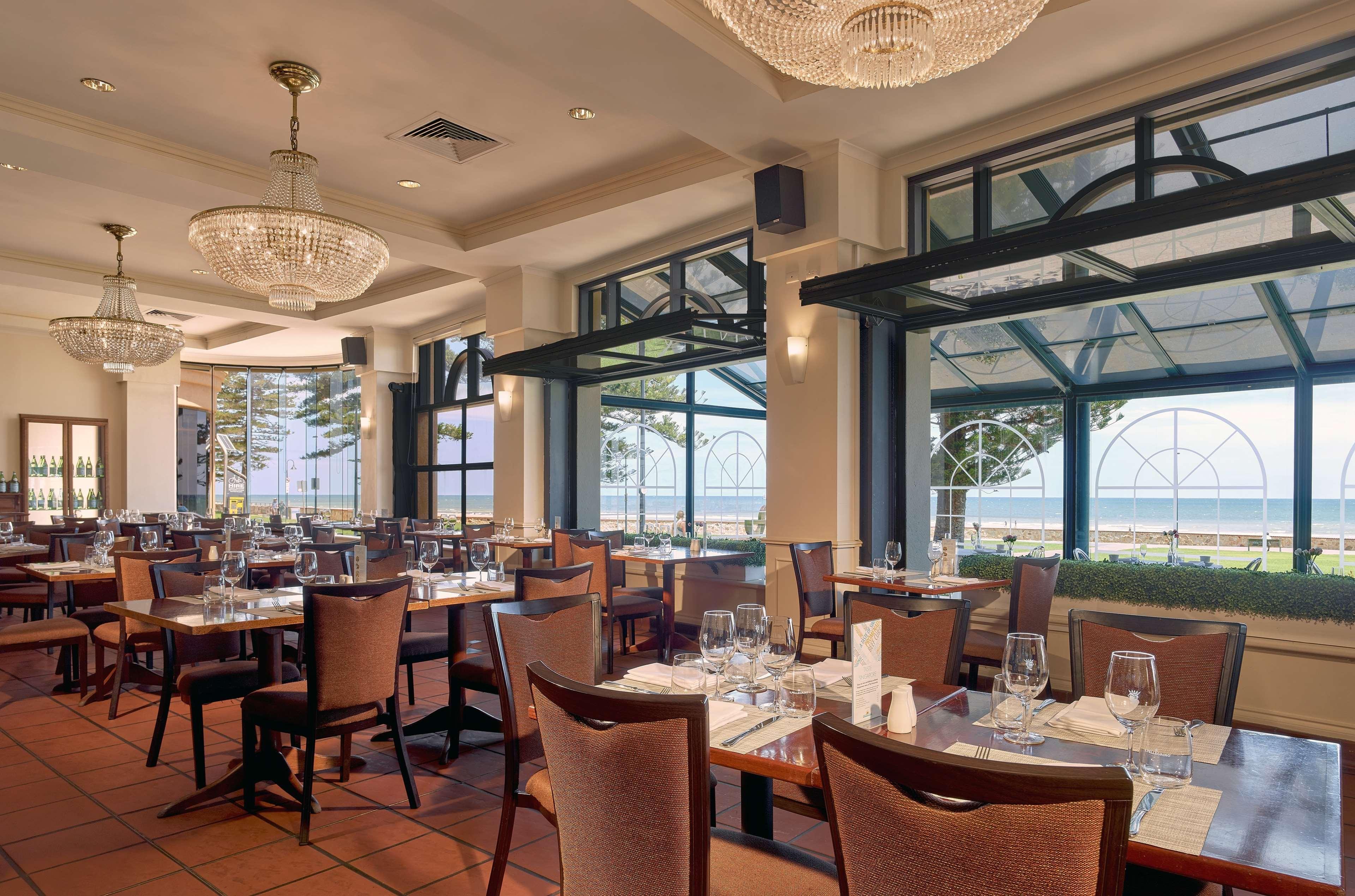 Hotel Stamford Grand Adelaide Glenelg Zewnętrze zdjęcie The photo shows the interior of a restaurant. It features several wooden tables set with glasses and utensils, suggesting it's ready for diners. The decor includes elegant chandeliers hanging from the ceiling, adding a touch of sophistication. Large 