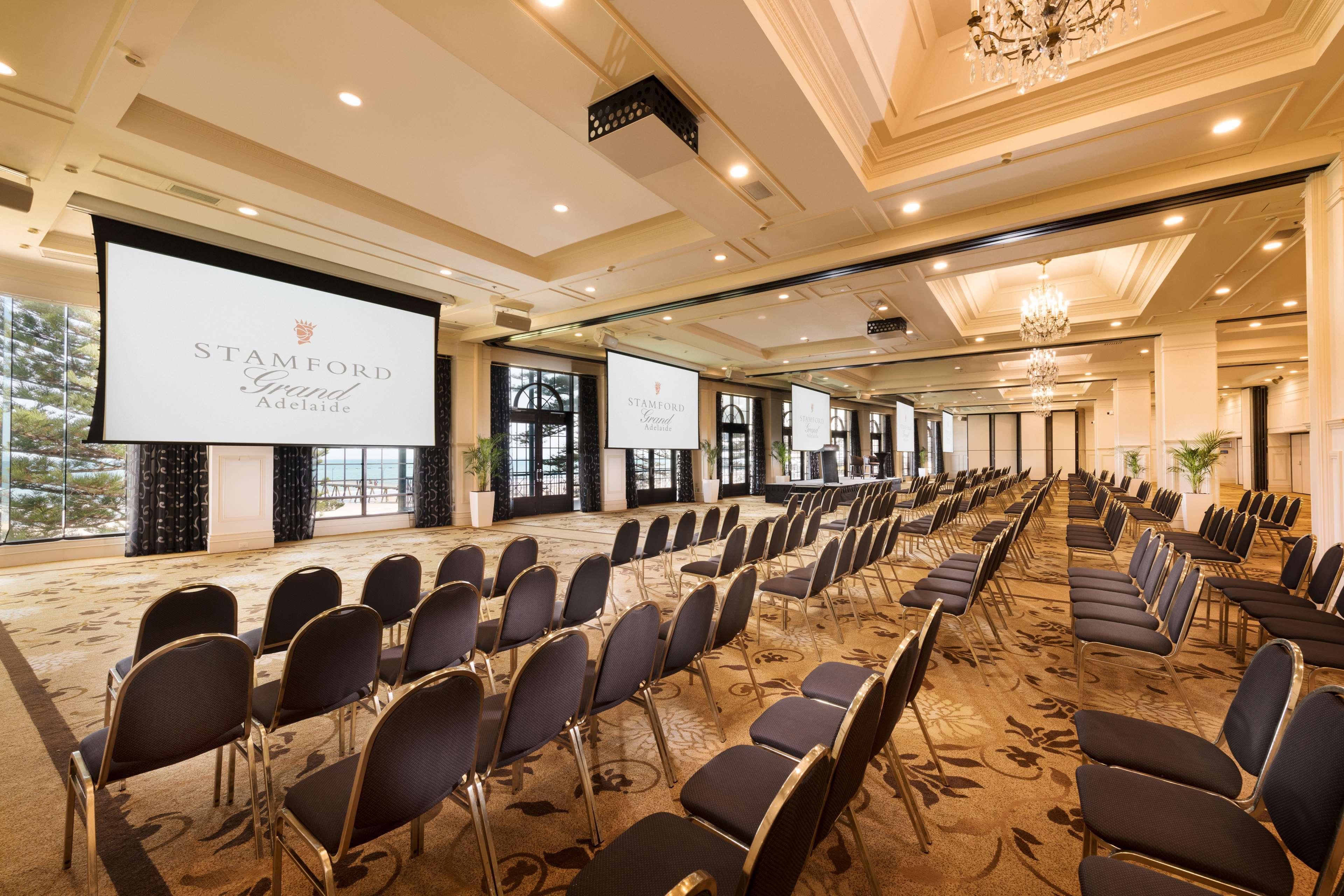 Hotel Stamford Grand Adelaide Glenelg Zewnętrze zdjęcie The photo shows a spacious event or conference room. There are rows of dark, upholstered chairs arranged facing two large screens. The room features elegant decor with soft lighting, a chandelier, and windows that allow natural light to filter in. Th