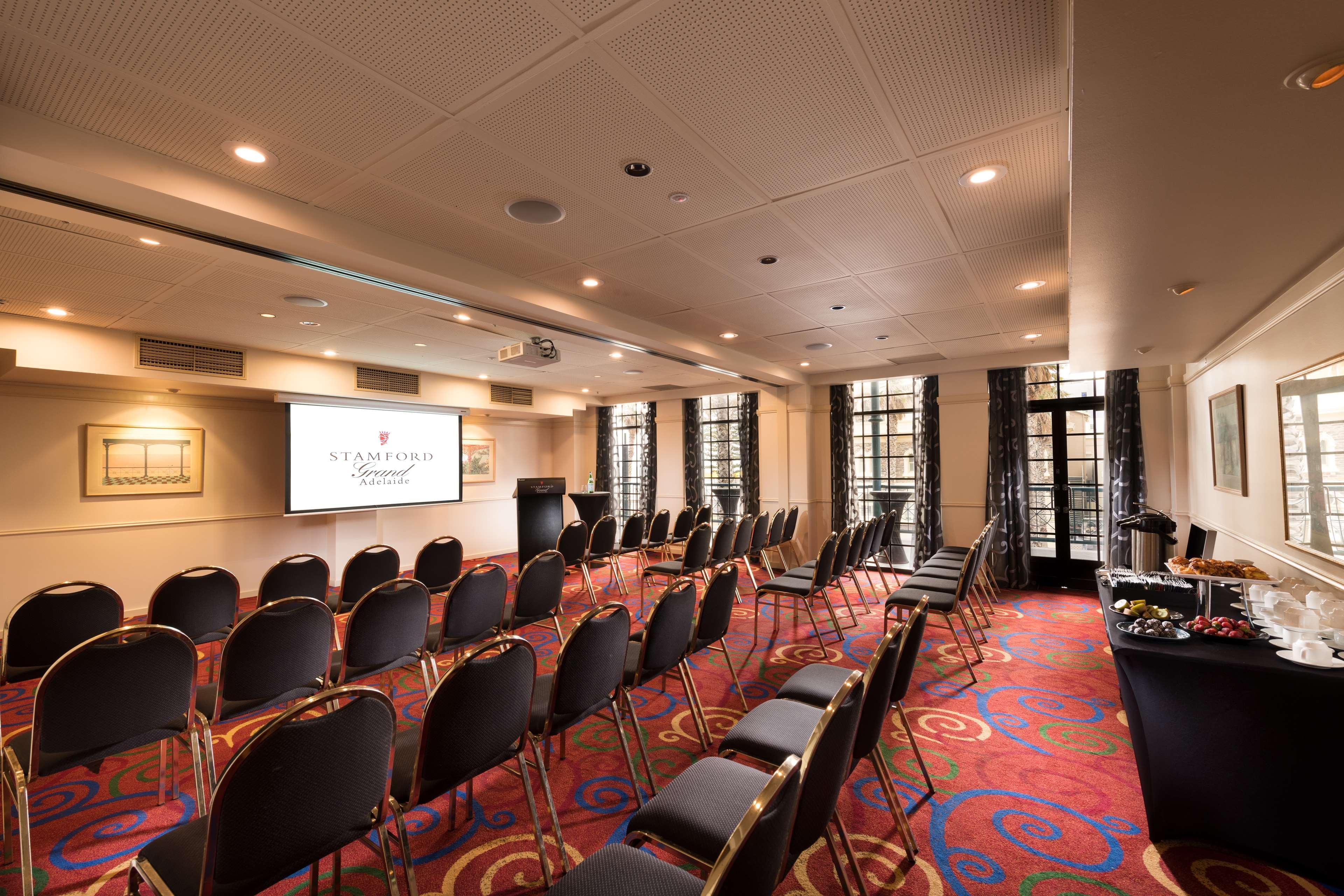 Hotel Stamford Grand Adelaide Glenelg Zewnętrze zdjęcie The photo shows a conference room set up for an event. There are rows of black chairs arranged in a semicircular formation facing a presentation screen at the front. The room features large windows that allow natural light to enter, and the flooring 