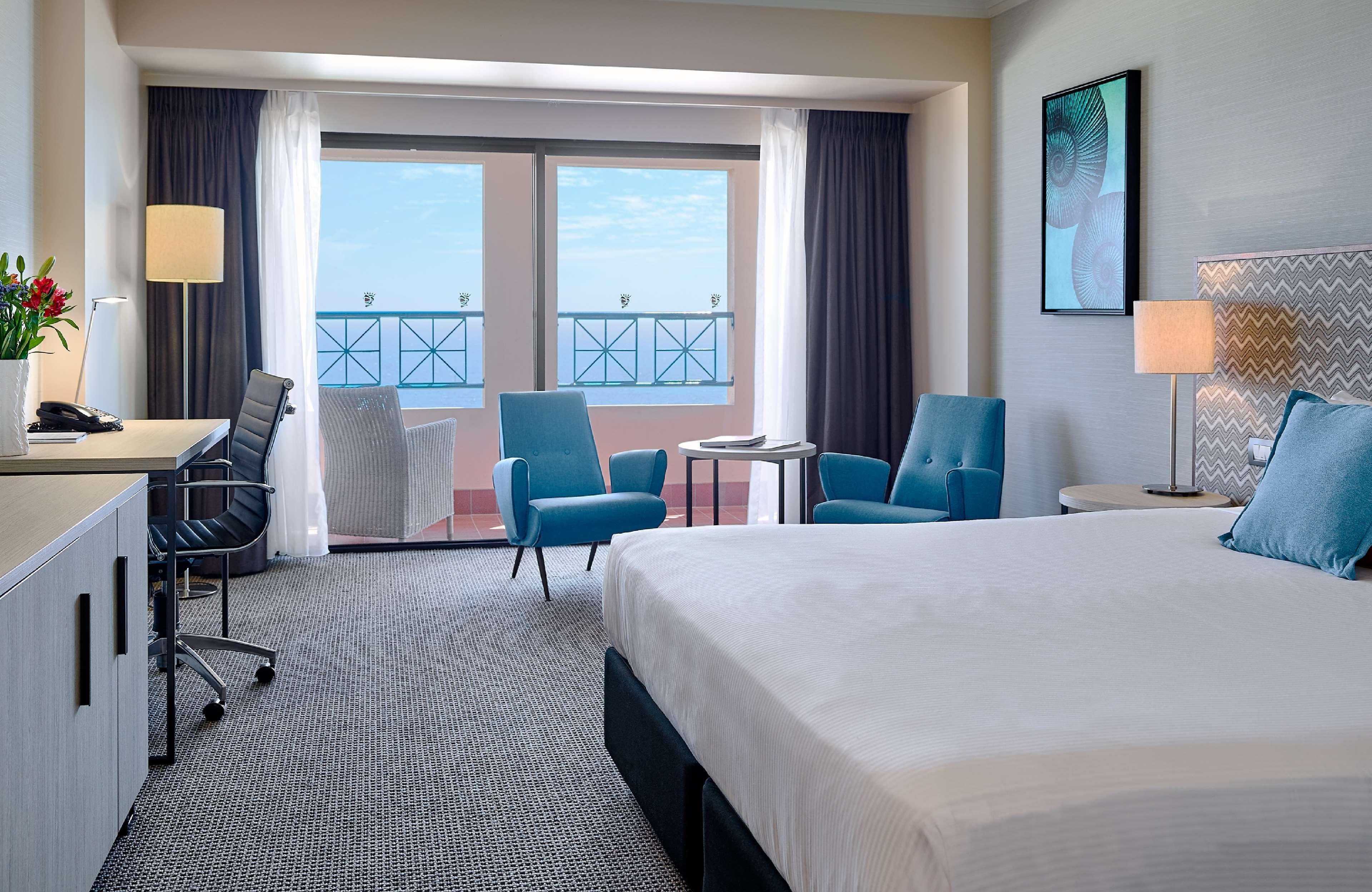 Hotel Stamford Grand Adelaide Glenelg Zewnętrze zdjęcie The image shows a hotel room featuring a large bed with a white duvet. There are two bright blue chairs and a small table near a large window that provides a view outside, possibly to a balcony. The room includes a workspace with a desk and an office