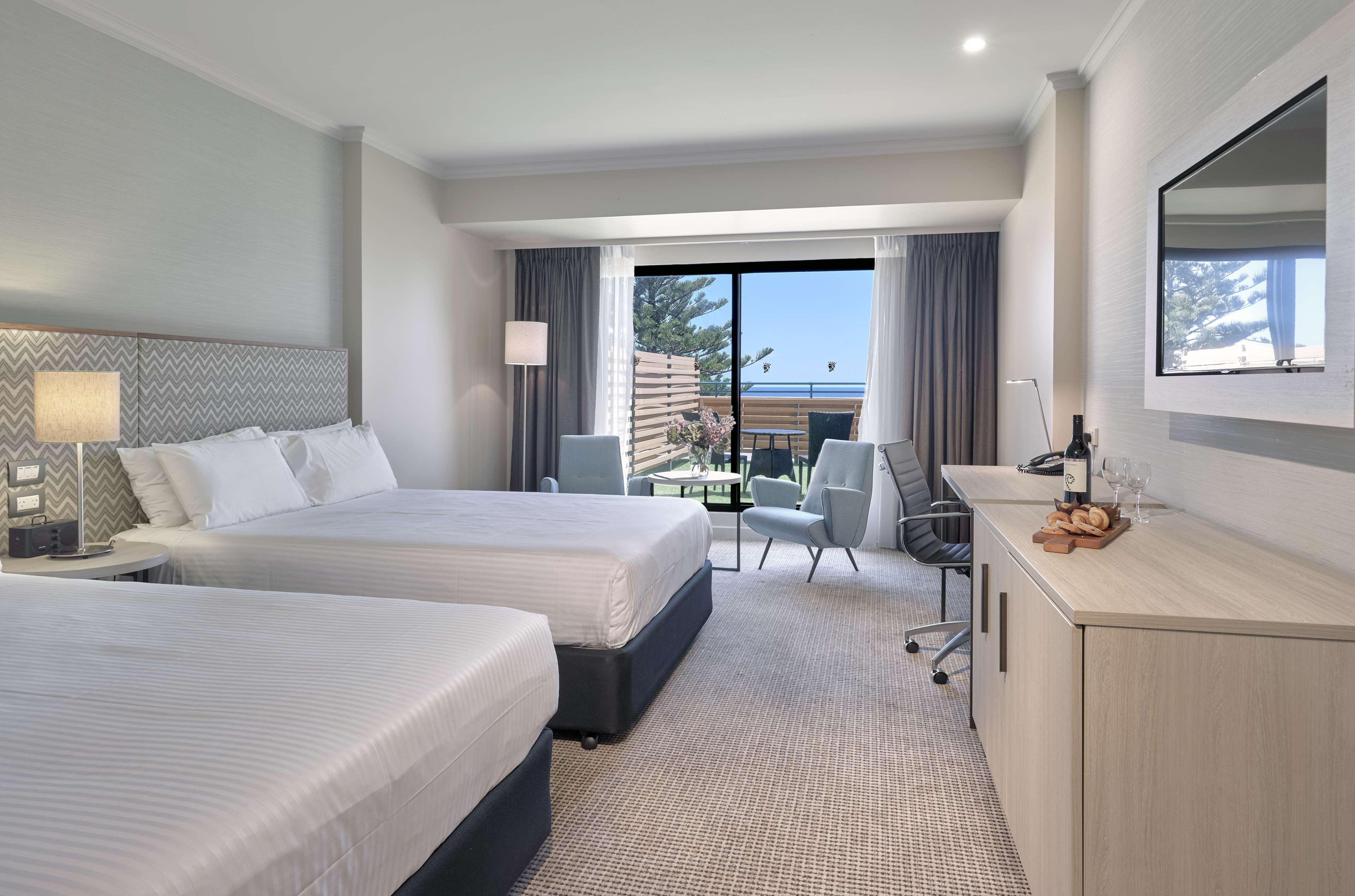 Hotel Stamford Grand Adelaide Glenelg Zewnętrze zdjęcie The photo shows a modern hotel room featuring two beds with white linens. There's a large window that provides natural light and a view of a balcony area. The room includes two chairs and a small table, creating a cozy sitting area. A television is m