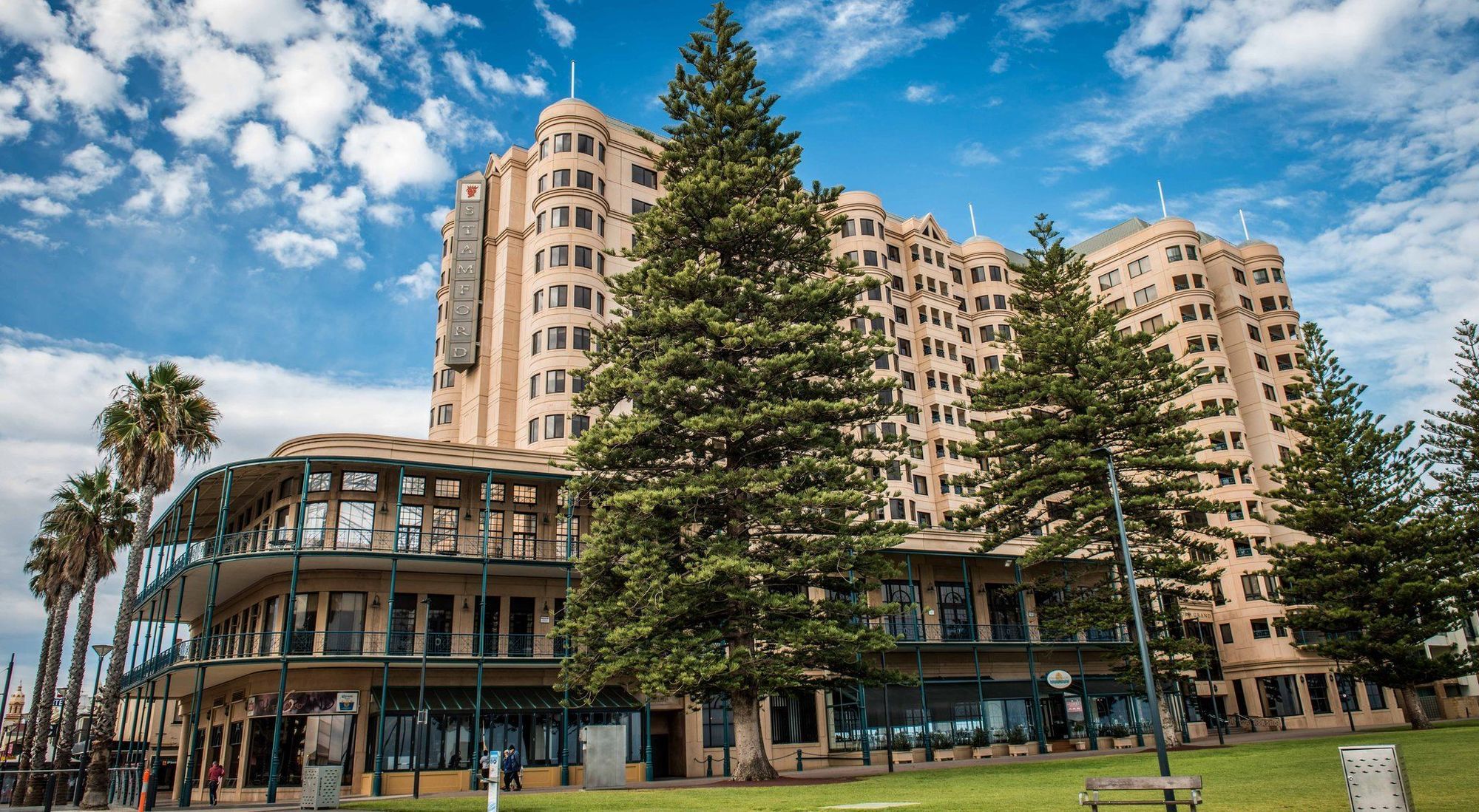 Hotel Stamford Grand Adelaide Glenelg Zewnętrze zdjęcie The image shows a large building with a distinctive architectural style, characterized by a tan or beige color. The structure appears to have multiple floors and rounded edges. In front of the building, there are several tall green trees, which add t